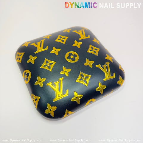 A square, dark green leather elbow cushion pad, made of PU leather material, showcases a pattern of yellow floral designs and stylized initials set against a white backdrop. The top left corner is adorned with colorful text that reads "Dynamic Nail Supply," making it an ideal addition to your manicure nail desk. Along the bottom edge, the website address repeats in white text.