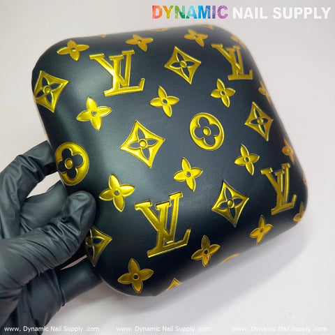 A gloved hand holds a square black Leather Elbow Cushion Pad Arm Rest Support for Manicure Nail Desk, crafted from PU leather and adorned with yellow monograms and floral motifs. The luxury brand design is embossed on the pad, set against a plain white background. At the top edge, the branding text "Dynamic Nail Supply" suggests its use as an elegant manicure nail desk accessory.