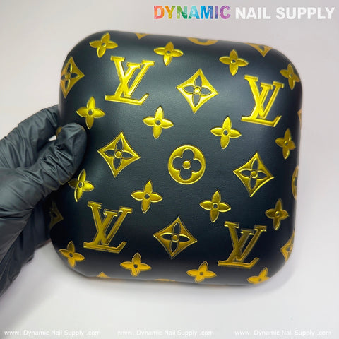 A gloved hand holds a square elbow cushion pad, crafted from PU leather with an intricate pattern of gold floral and geometric motifs. At the top of the background is text saying Dynamic Nail Supply, making it perfect for adding elegance to any manicure nail desk setup.