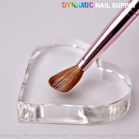 The Dynamic Metallic Pink Kolinsky Hair Brush rests on a clear heart-shaped glass dish atop a white surface.