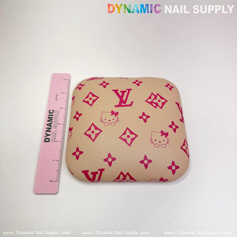 Add a touch of style to your manicure desk with the Khaki LV+Kitty Pattern Leather Elbow Cushion Pad Arm Rest Support from Dynamic Nail Supply. This square, beige cushion features pink Hello Kitty and LV patterns, providing excellent support for your arms. It also includes a pink ruler labeled "DYNAMIC" on the left side for scale, all set against a plain white background.