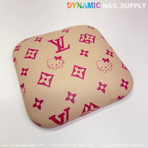 The Khaki LV+Kitty Pattern leather elbow cushion pad for a manicure nail desk serves as an armrest support, embellished with pink designs that include floral motifs, bow-adorned cat faces, and stylized letters. The words "Dynamic Nail Supply" are displayed in the top right corner, with the company website written along the bottom edge in white text.