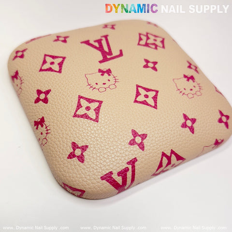 A square, khaki elbow cushion pad featuring a leather surface with a red Hello Kitty and geometric design reminiscent of luxury branding provides ideal support at a manicure nail desk. The brand "Dynamic Nail Supply" is prominently displayed in colorful letters above, with the website www.DynamicNailSupply.com along the bottom edge.