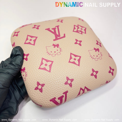 A gloved hand holds a square khaki cushion, adorned with a pink pattern featuring Hello Kitty faces and floral motifs. This Leather Elbow Cushion Pad from Dynamic Nail Supply provides perfect arm rest support for your manicure desk. The plain white background accentuates the textured surface and the colorful logo of Dynamic Nail Supply along the top edge.