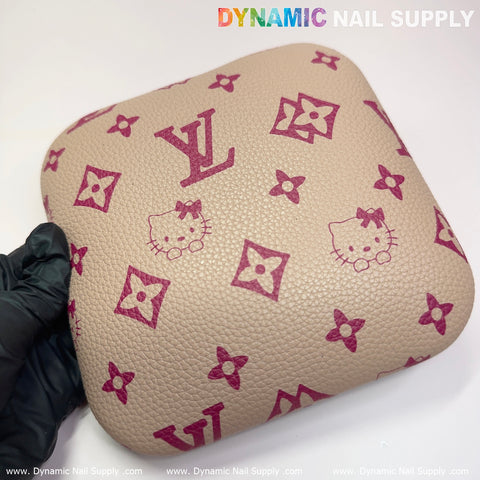 A square khaki object with a textured surface features pink designs, including a cartoon cat face and floral motifs. A gloved hand holds the object resembling an elbow cushion pad, ideal for manicure desk setups. The text Dynamic Nail Supply is visible at the top and bottom edges.