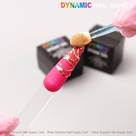 A gloved hand uses No-wipe Ready for Chrome Spider Gel to create gold lines on pink, gold-swirled nail tips.