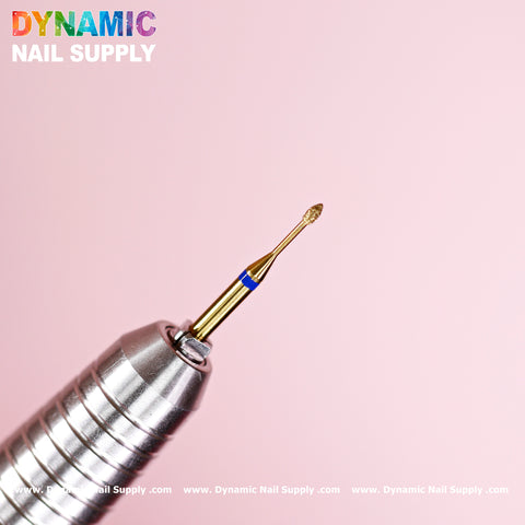 A close-up of the Medium Fine Cuticle Drill Bit (Tiny Ellipse Head) by Dynamic Nail Supply, featuring a gold finish against a pink background. Designed for Russian nail manicure and cleaning the cuticle area, it has a blue band near its base connected to a metallic, ridged handle. In the top left corner is the vibrant logo of Dynamic Nail Supply.