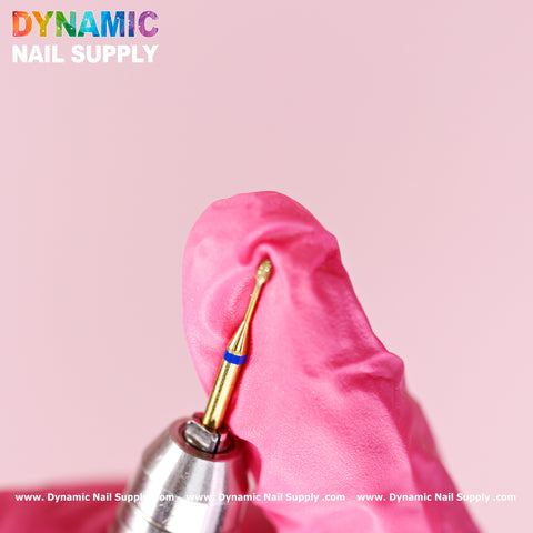 A gloved hand holds the Medium Fine Cuticle Drill Bit (Tiny Ellipse Head) against a pink background. Designed for precision in Russian nail manicures, the metallic bit features yellow and blue rings near the base. Bold rainbow text reads "Dynamic Nail Supply," with a website link in white at the bottom.