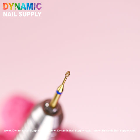 Close-up of the Dynamic Nail Supply Medium Fine Cuticle Drill Bit (Tiny Ellipse Head) on a soft pink background. Perfect for Russian Nail Manicure, it is displayed in a metallic holder. The vibrant Dynamic Nail Supply logo is prominently at the top, with their website text repeated horizontally at the bottom.