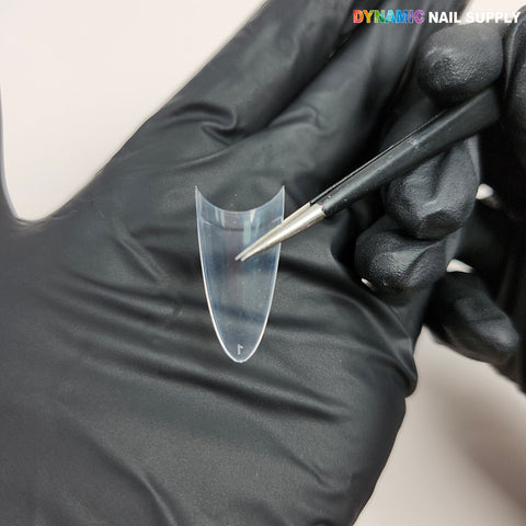 A gloved hand holds a Medium Almond Extension Nail Tip as a brush touches it; DYNAMIC NAIL SUPPLY is in the corner.