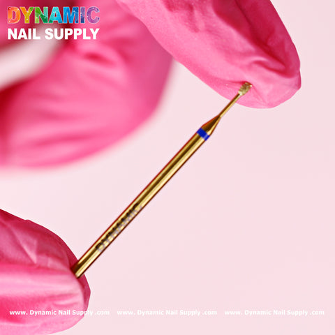 A close-up shows a hand adorned with a pink glove holding the Medium Fine Cuticle Drill Bit (Tiny Ellipse Head) with a blue stripe near its base, perfect for Russian Nail Manicure and cuticle cleaning. The backdrop is light pink, featuring the Dynamic Nail Supply logo in vibrant lettering at the top, while the website URL appears repeatedly at the bottom.