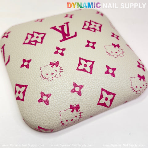 A square, textured leather armrest pad adorned with pink Hello Kitty faces and floral motifs evocative of a well-known brand serves as a stylish elbow cushion. The beige background enhances the playful design, with "Dynamic Nail Supply" subtly visible along the edges. Ideal for any manicure nail desk.