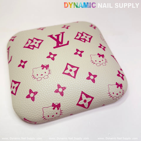 Introducing the Beige LV+Kitty Pattern Leather Elbow Cushion Pad from Dynamic Nail Supply. This cream-colored cushion, adorned with pink designs featuring a stylized LV logo, Hello Kitty faces, and floral-like symbols, is ideal for arm rest support at your manicure nail desk. Its pebbled texture provides comfort and style, while the upper right corner showcases "DYNAMIC NAIL SUPPLY" in bold, multicolored text.