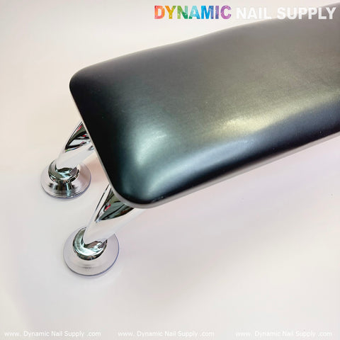 A black microfiber leather armrest with a cushion pillow for pedicure and manicure services, featuring shiny silver chrome legs, is positioned diagonally on a white surface. At the top, the brand name Dynamic Nail Supply is displayed in colorful lettering, paired with an anti-slip bracket to ensure stability during manicure sessions.