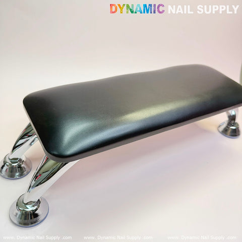 A black microfiber leather armrest with a cushion pillow designed for pedicures and manicures, featuring a sleek rectangular shape, rests securely on an anti-slip bracket supported by four shiny metallic legs, centered on a white background. The brand name, Dynamic Nail Supply, is printed in colorful text in the upper right corner, with the website URL repeating along the bottom edge.