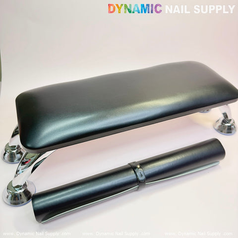 A black Microfiber Leather Armrest with a cushion pillow for pedicures and manicures from Dynamic Nail Supply sits elegantly on a white surface, supported by chrome legs. Below it, a rolled-up black mat is neatly secured with a strap. Ideal for any manicure service, this setup is enhanced by the Dynamic Nail Supply watermark repeated in the background.