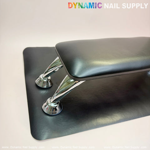 A stylish black microfiber leather armrest features two polished chrome supports and is accompanied by an anti-slip bracket on a coordinating black mat. Its surface is smooth and lightly cushioned, with "Dynamic Nail Supply" inscribed at the top right corner.