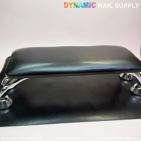 A black microfiber leather armrest with a cushioned pillow and chrome legs rests on a matching mat, equipped with an anti-slip bracket for added stability. The Dynamic Nail Supply logo is displayed in vibrant gradient text on a white background that accentuates the sleek design, making it ideal for pedicure and manicure services.