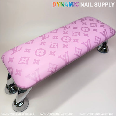 The "Big Logo Pink LV Pattern" Leather Armrest Cushion Pillow for Pedicure Manicure, ideal for any nail service, showcases a stylish design with interlocking symbols and letters. It features four shiny, curved metal legs set against a neutral gradient background. The brand name "Dynamic Nail Supply" is displayed in colorful letters at both the top and bottom edges.