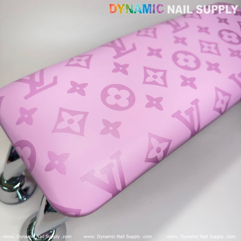 A pink leather armrest cushion, featuring an LV pattern of stylized floral and star shapes reminiscent of a luxury brand, is supported by shiny metallic legs. The Dynamic Nail Supply logo is partially visible at the top, making it perfect for elevating your manicure nails service aesthetic.