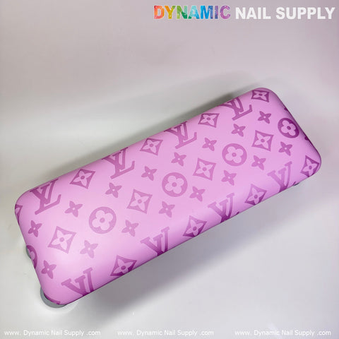 A rectangular armrest cushion, featuring a pink leather LV pattern with white floral accents, provides an ideal support for your manicure session. The brand name "Dynamic Nail Supply" is displayed at the top, while "www.DynamicNailSupply.com" is repeated along the bottom. This product is from Dynamic Nail Supply's collection of manicure accessories.