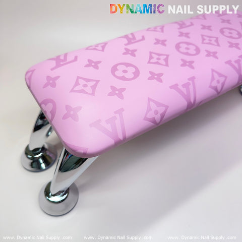 The Big Logo Pink LV Pattern Leather Armrest Cushion Pillow for Pedicure Manicure by Dynamic Nail Supply is perfect for any manicure service. It showcases a stylized symbol pattern and rests on shiny chrome legs, with "DYNAMIC NAIL SUPPLY" displayed in colorful, bold letters above. The clean and modern design makes it ideal for salons using a UV nail light.