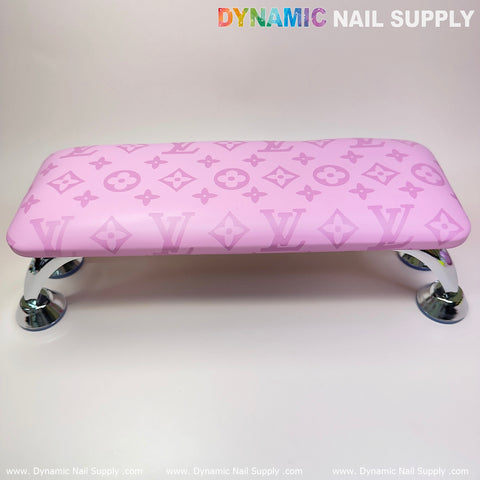 The (Big Logo Pink LV Pattern) Leather Armrest Cushion Pillow for Pedicure Manicure from Dynamic Nail Supply is showcased, featuring pink cushioning with a stylish pattern of symbols and letters and supported by chrome legs, making it ideal for a manicure service. The clear white background includes the Dynamic Nail Supply text in gradient colors at the top corners.