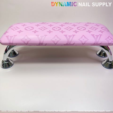 A pink leather armrest cushion pillow featuring the LV pattern design rests on four shiny silver metal legs, perfect for any manicure service. It stands against a white background with "Dynamic Nail Supply" written in colorful and gray letters at the top, while "www.DynamicNailSupply.com" repeats along the bottom edge.