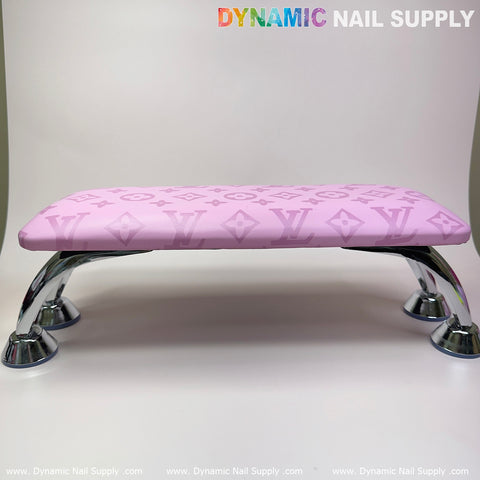 A pink leather armrest cushion pillow for pedicures and manicures, branded by Dynamic Nail Supply, is adorned with an LV pattern of interlocking letters and diamond shapes. It rests elegantly atop four gleaming metal legs against a plain white backdrop, with "Dynamic Nail Supply" prominently displayed in the top right corner.