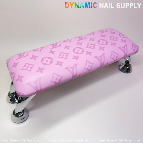 The "(Big Logo Pink LV Pattern) Leather Armrest Cushion Pillow for Pedicure Manicure" by Dynamic Nail Supply features a floral motif with stylized LV letters on pink leather and chrome legs. The top left corner prominently displays "DYNAMIC NAIL SUPPLY" in multicolored font, while its base elegantly reflects on a glossy white surface, making it an ideal accent for any manicure nails service.