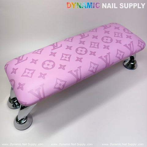 Introducing the Dynamic Nail Supply Leather Armrest Cushion Pillow for Pedicure and Manicure, in a chic pink LV pattern. This stylish accessory boasts floral and letter motifs, along with vibrant brand lettering on top. It comes equipped with sleek metal legs and rests on a reflective white surface, making it the perfect addition to any manicure nail service setup.