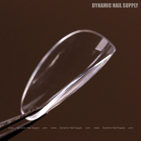 A Natural Almond (Short) Full Cover Soft-Gel Nail Tip is held by dark tweezers against a brown background.