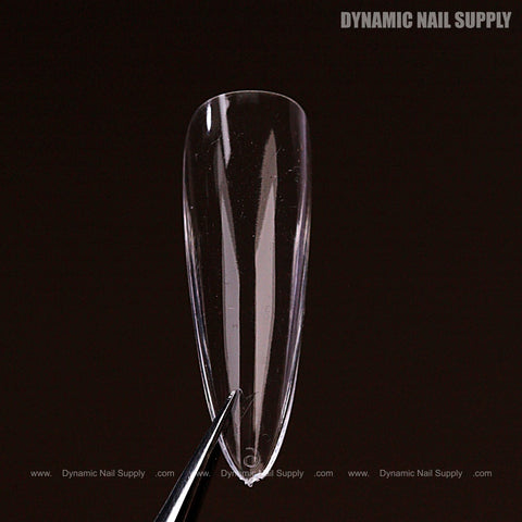 A Natural Almond (Long) Full Cover Soft-Gel Nail Tip (Pink Box) held by tweezers against a dark background.