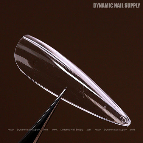 A Natural Almond (Long) Soft-Gel Nail Tip from the Pink Box held by silver tweezers against a dark brown background.