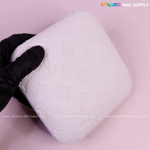 A gloved hand holds a White LV Engrave-Grain Elbow Rest Pad for Nail Tech on a pink background.