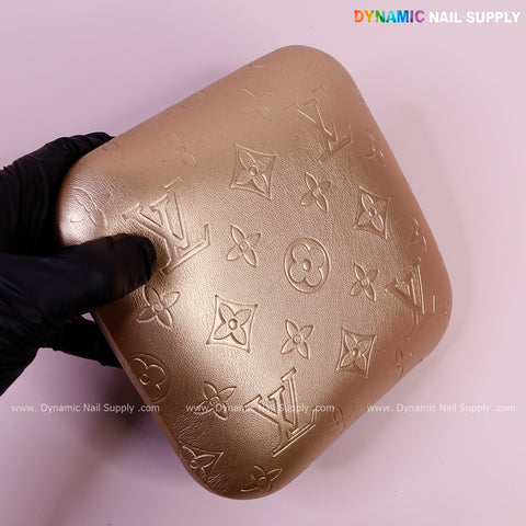 A gloved hand holds the Metallic Gold LV Engrave Elbow Rest Pillow for Nail Tech on a light pink branded surface.