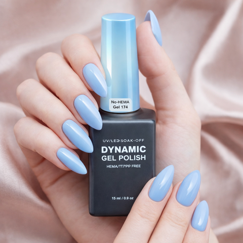 A hand with glossy light blue nails holds a Dynamic No-HEMA Gel Polish (Color 171-180) on satin fabric.