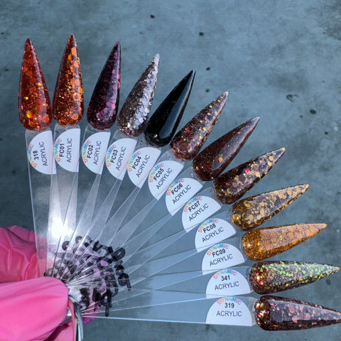 A fan display of 12 pointed acrylic nail samples, held by someone wearing a pink glove, showcases the "Flavors Of Fall" 2024 Acrylic Powder Collection by Dynamic Nail Supply. Featuring glitter shades of red, orange, brown, black, and gold, these non-toxic acrylic nails are crafted to last and each is labeled with its color number. The background is set against a textured gray surface.