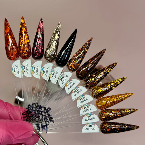 A hand adorned with a pink glove holds an array of elongated, pointed acrylic nail samples created using the non-toxic "Flavors Of Fall" 2024 Acrylic Powder Collection (part 4) Newest by Dynamic Nail Supply. The vibrant nails showcase a diverse palette of colors and shimmering glitter patterns, including hues of red, pink, gold, black, and silver. Each sample is identified by a code against a neutral beige background.