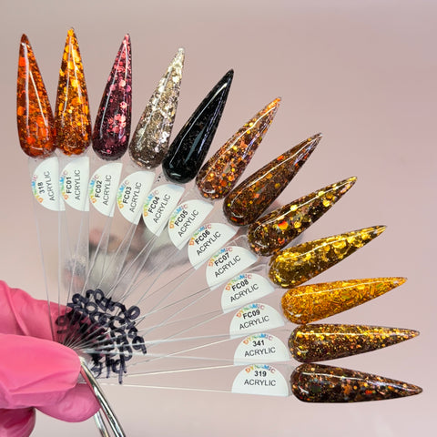 A hand wearing a pink glove holds a fanned set of long, stiletto-shaped nail samples from the "Flavors Of Fall" 2024 Acrylic Powder Collection by Dynamic Nail Supply. These non-toxic nails showcase glitter designs with multicolored flecks and gold sparkles. Each sample is labeled with a code and the collection name, all resting against a soft, muted pink background.