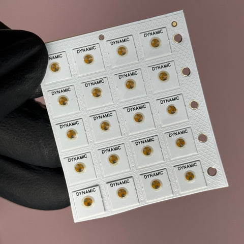 A gloved hand holds a small white sheet featuring several NFC Tag Nail Chips from Dynamic Nail Supply, each displaying a metallic circular element in the center. These microchips, branded as DYNAMIC, are smartphone readable and designed for viral mobile marketing. The soft pinkish hue in the background enhances the appeal of this innovative product.