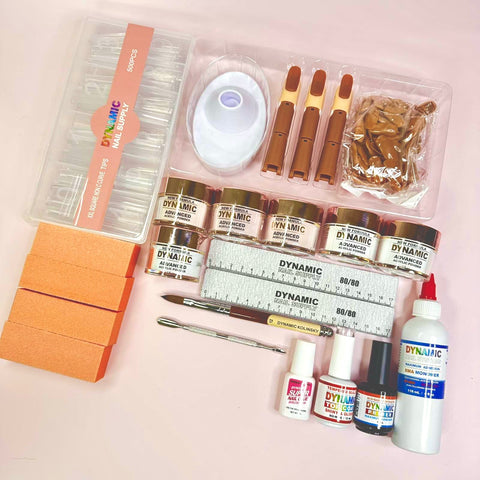 The Dynamic Nail Supply Acrylic Nail Starter Kit for professionals and beginners is displayed like a professional nail salon setup, featuring a variety of tools and supplies. Neatly arranged on a light pink surface are clear nail tips in a container, several jars of color powders, files, buffers, nail glue, a sculpting tool, and a bottle of liquid monomer.