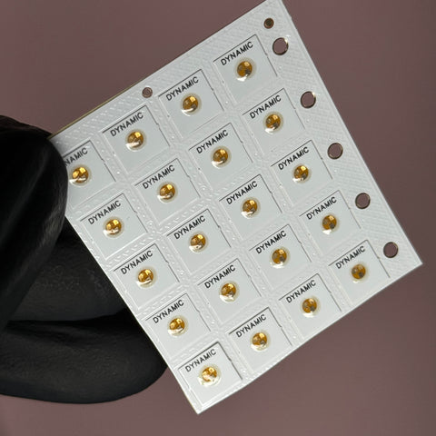 A gloved hand holds a micro-sized grid of NFC Tag Nail Chips, each labeled with "Dynamic Nail Supply" and featuring a central gold dot. Perforations on the right side allow for attachment or separation. The soft, out-of-focus brownish background accentuates the intricate design, making it perfect for smartphone-readable applications in viral mobile marketing.