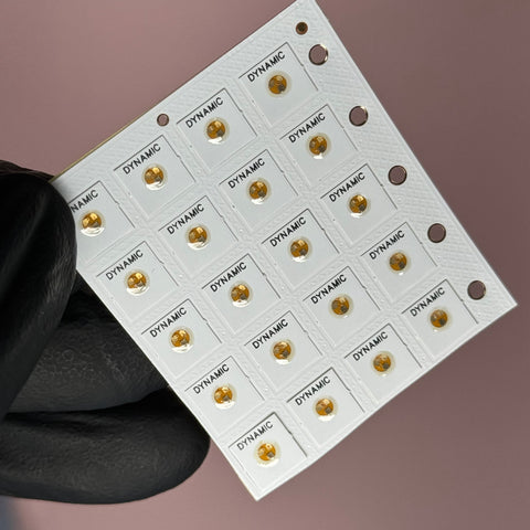 A gloved hand holds a small white perforated grid featuring multiple rectangular labels. Each label, adorned with the Dynamic Nail Supply's NFC Tag Nail Chips logo in black, includes a small circular gold dot that evokes the appearance of a micro-sized chip, all set against a muted pink background.