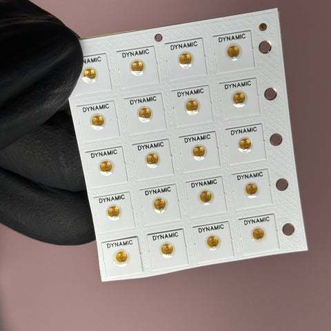 A gloved hand holds a small NFC Tag Nail Chip from Dynamic Nail Supply, featuring a grid of 20 square sections with each section showcasing a round, gold-colored component labeled DYNAMIC. This smartphone-readable microchip includes a line of tiny holes on the right edge. The background is a soft pink.