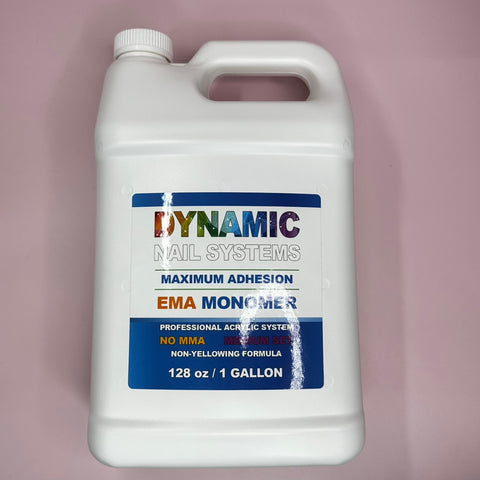 A white gallon container from Dynamic Nail Supply rests on a pink surface, labeled Dynamic Nail System Monomer - EMA Nail Liquid - Healthier Nail Liquid Monomer. The label highlights features such as Maximum Adhesion, No MMA, Medium Set time, and a Non-Yellowing Formula. Enhanced with color stabilizers, this 128 oz / 1-gallon nail liquid monomer is both vibrant and striking.