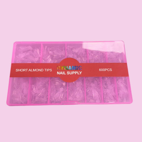 A pink organizer box with a transparent lid contains Short Almond Nail Tips - Half Cover, made from flexible material and arranged in multiple compartments. A red label on the box reads DYNAMIC NAIL SUPPLY, SHORT ALMOND TIPS, 600 PCS. The background is light pink.