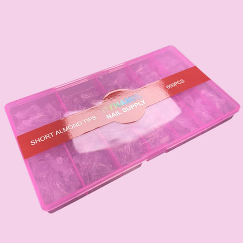 A pink plastic container from Dynamic Nail Supply, labeled "Short Almond Nail Tips - Half Cover - 600 pcs 12 sizes (Short Stiletto)," features clear acrylic nail tips made from flexible material, neatly organized in compartments. The light pink background complements the collection of versatile almond nail tips.