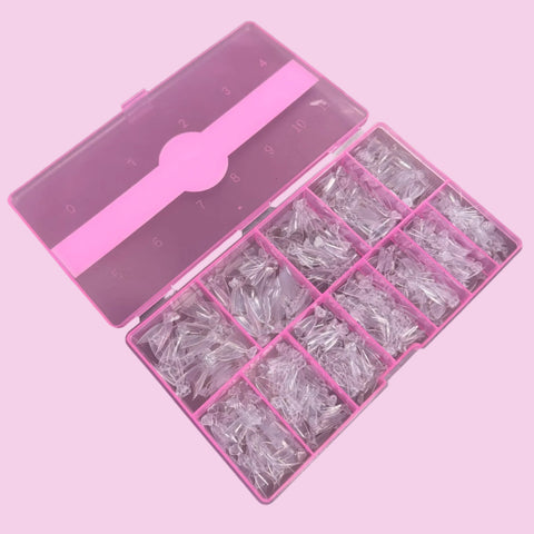 The Dynamic Nail Supply's Short Almond Nail Tips feature transparent, disposable short stiletto tips displayed in a stylish pink plastic organizer box. This set, which is divided into 12 labeled compartments for various sizes neatly arranged in rows, is made from flexible material and perfectly matches the pink background.