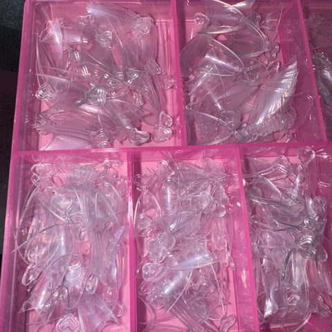 A pink plastic organizer from Dynamic Nail Supply holds several compartments filled with their Short Almond Nail Tips, each made of clear, glossy plastic shaped like butterflies. The sections are neatly arranged, showcasing a shiny surface that reflects light. These flexible nail tips are reminiscent of short stiletto shapes, adding an elegant touch to the display.